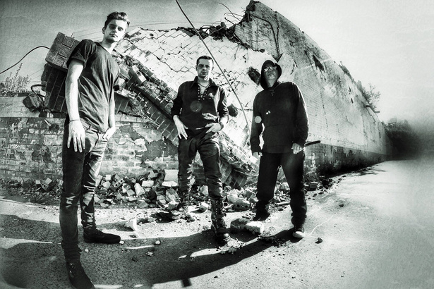 a black-and-white photo of three members of band shadowplay standing in front of a brick wall